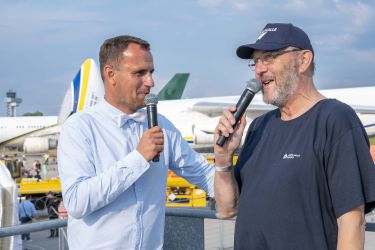 Zwei Männer mit Mikrofonen in der Hand, im Hintergrund Flugzeuge