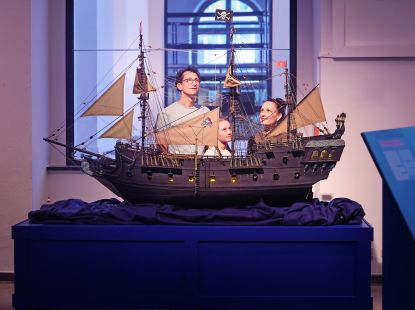 Eine Frau, ein Mann und ein Kind hinter einem großen Modell eines Segelschiffs mit Piratenflaggen
