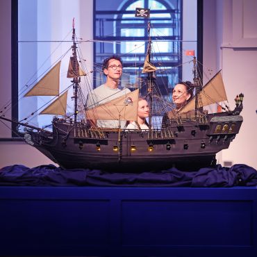 Eine Frau, ein Mann und ein Kind hinter einem großen Modell eines Segelschiffs mit Piratenflaggen