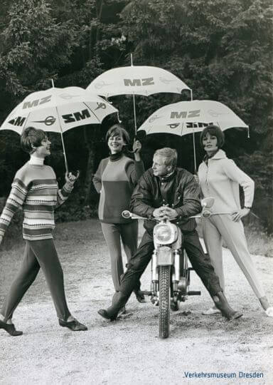 Drei Frauen mit einem Regenschirm stehen um ein Mann der auf einer MZ Trophy sitzt (Altes Werbefoto)